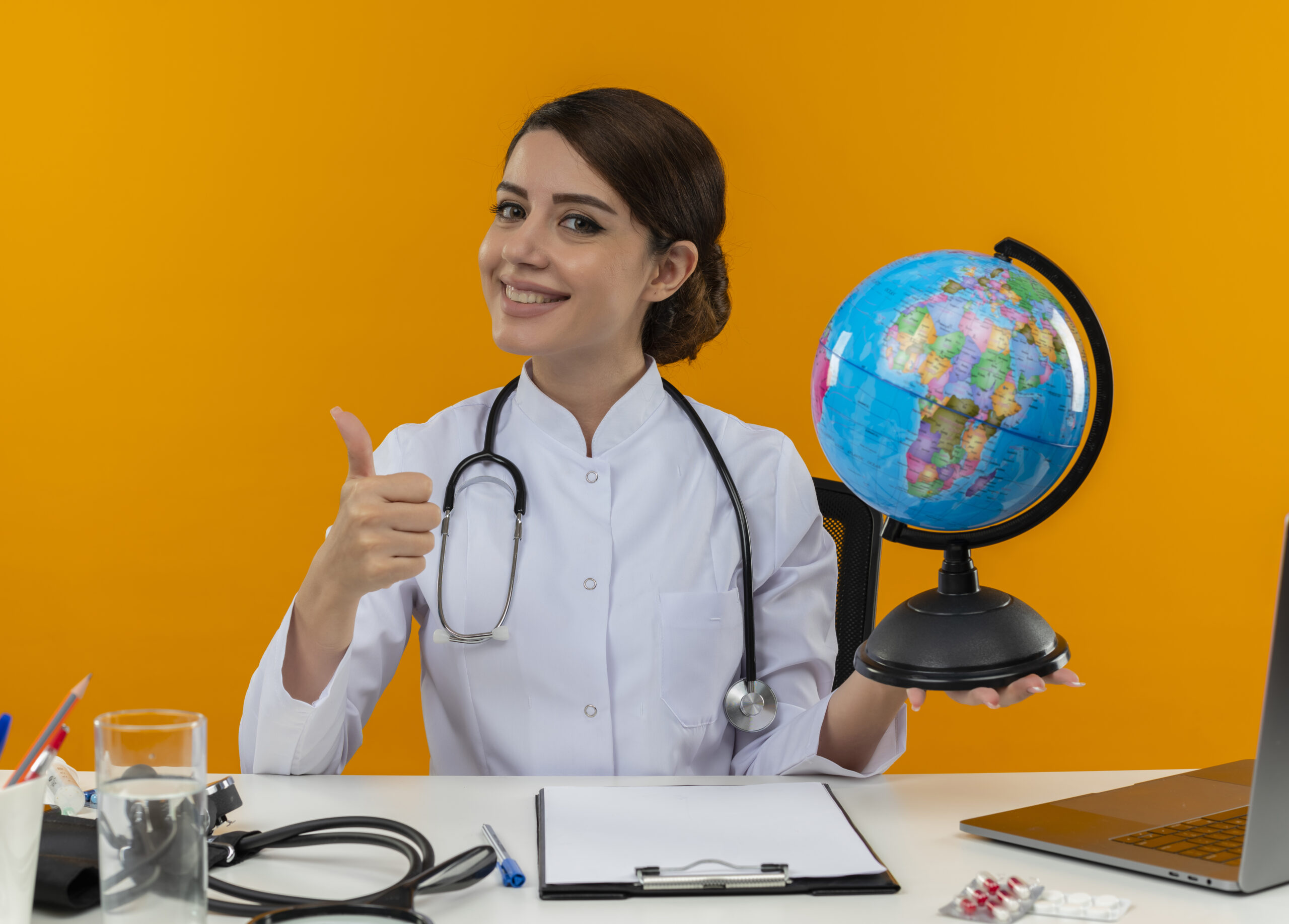 smiling young female doctor wearing medical robe and stethoscope sitting at desk with medical tools and laptop holding globe showing thumb up isolated on yellow background